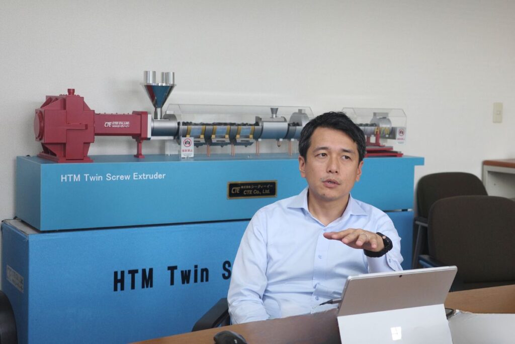 Masato Ito, CEO of CTE, discussing advanced extrusion technology with a model of the HTM Twin Screw Extruder in the background.