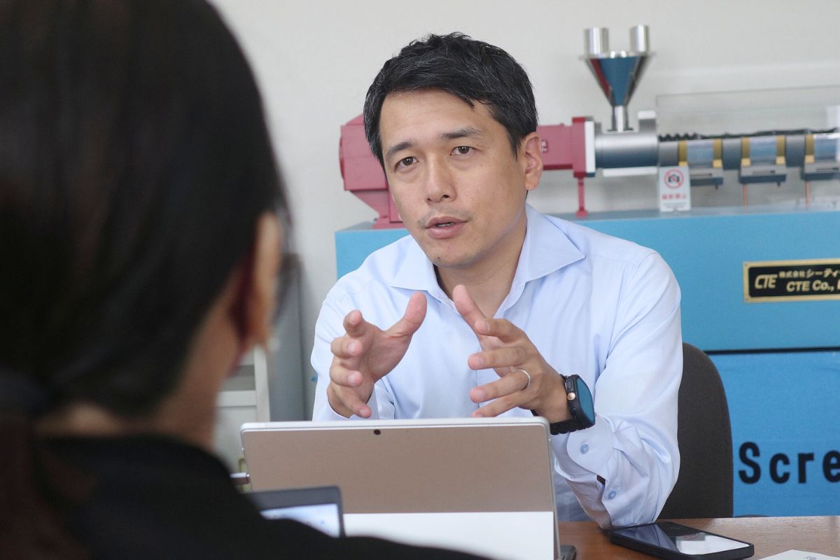 Masato Ito, CEO of CTE, engaging in conversation about extrusion technology with a model of the HTM Twin Screw Extruder visible behind him.