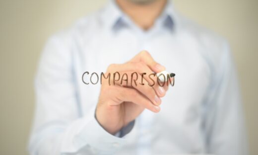 「COMPARISON」と書かれた透明なボードにマーカーで文字を書くビジネスマンの手元。比較や分析をテーマにしたコンセプトイメージ。